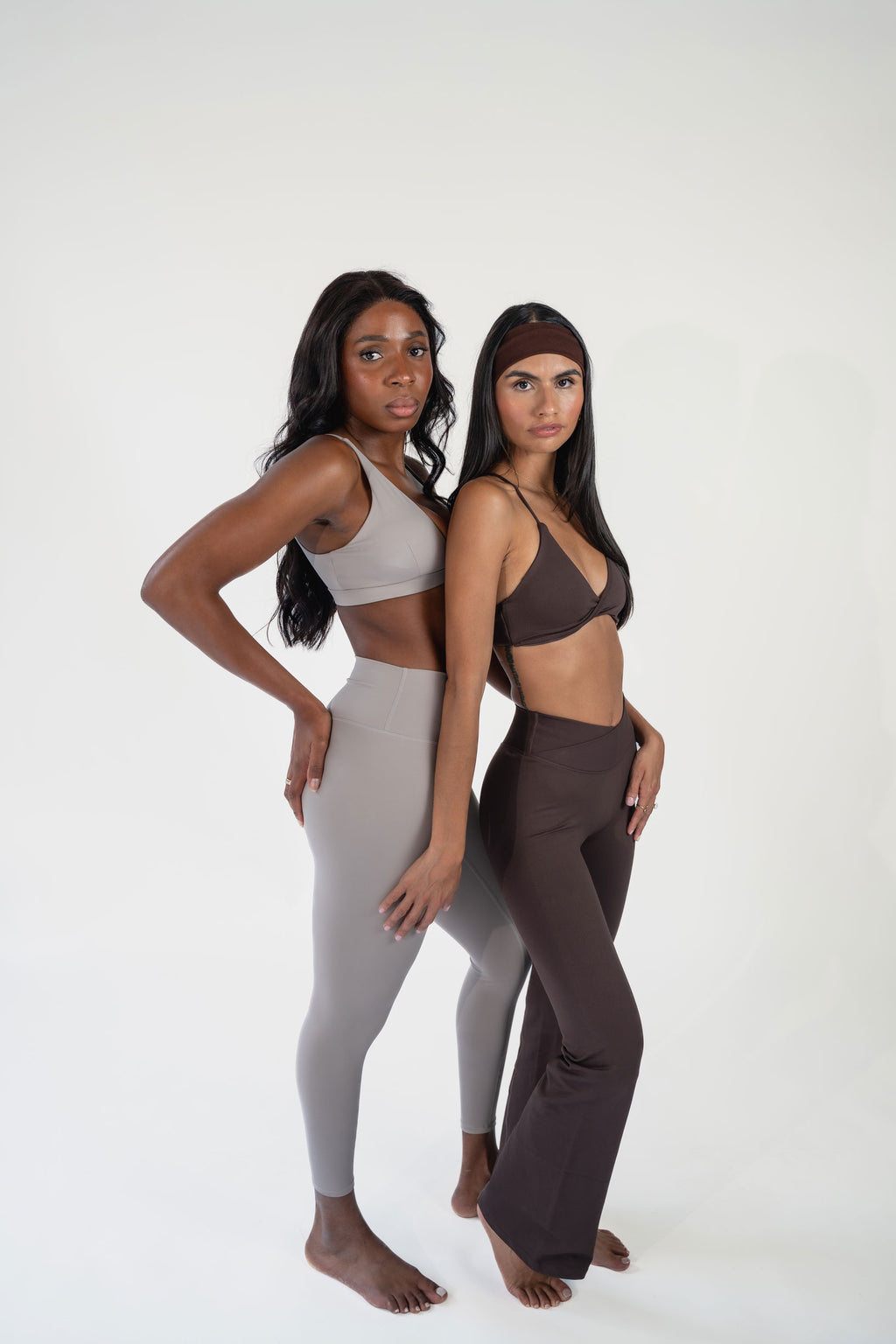 Two women modeling matching activewear sets against a plain white studio background. The woman on the left wears a light gray sports bra and leggings set, and the woman on the right wears a dark brown sports bra and matching pants with a brown headband.